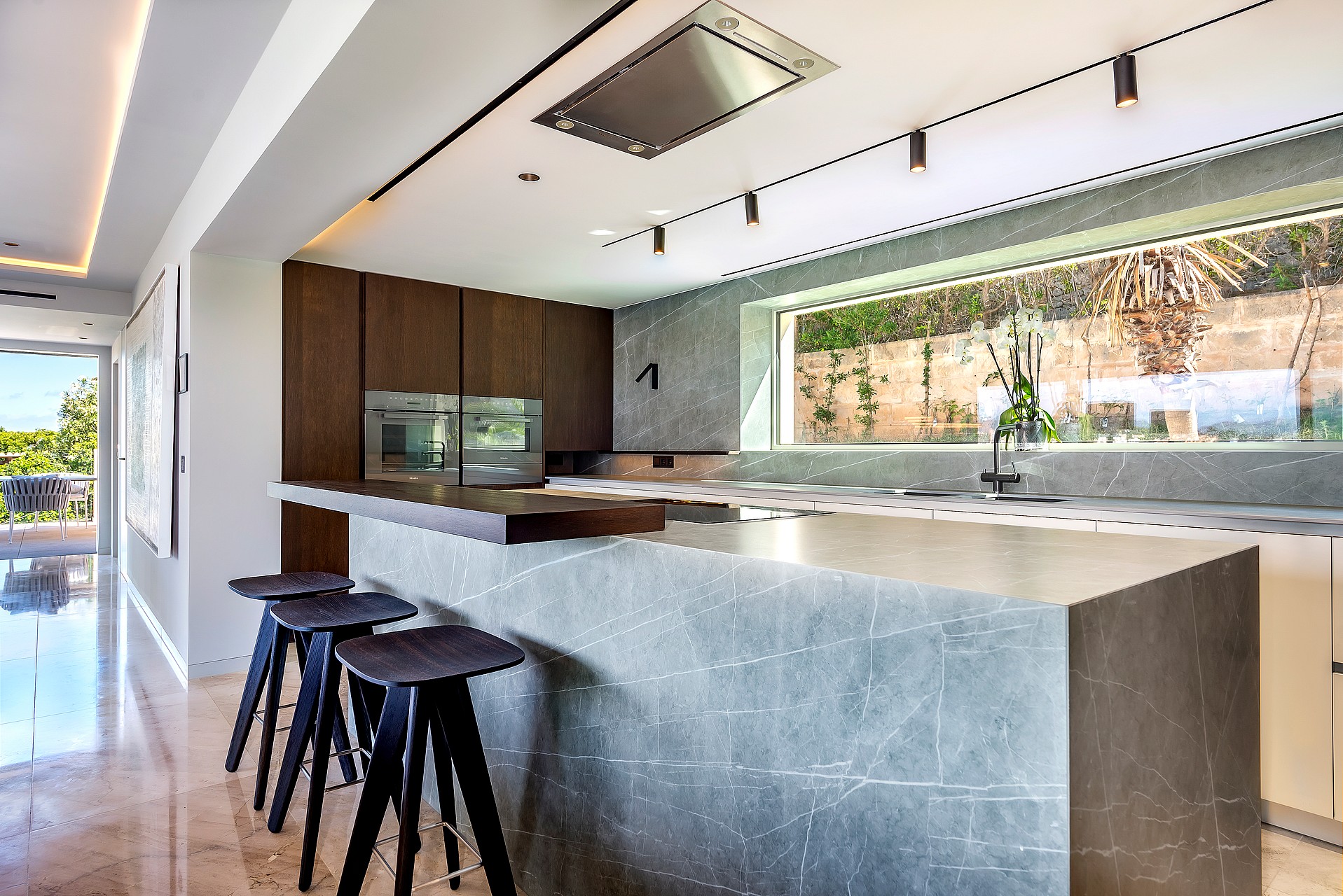 Modern kitchen with a large grey marble island, wooden cabinets, and three dark wooden bar stools under track lighting.