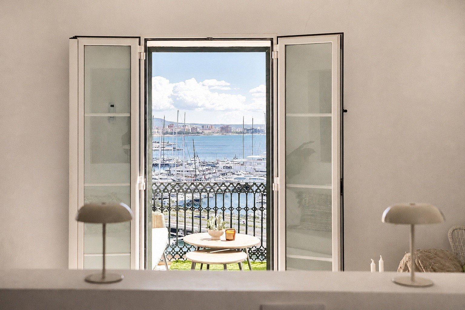 Interior view through open French doors to a small wrought-iron balcony overlooking a harbour with boats and city skyline.