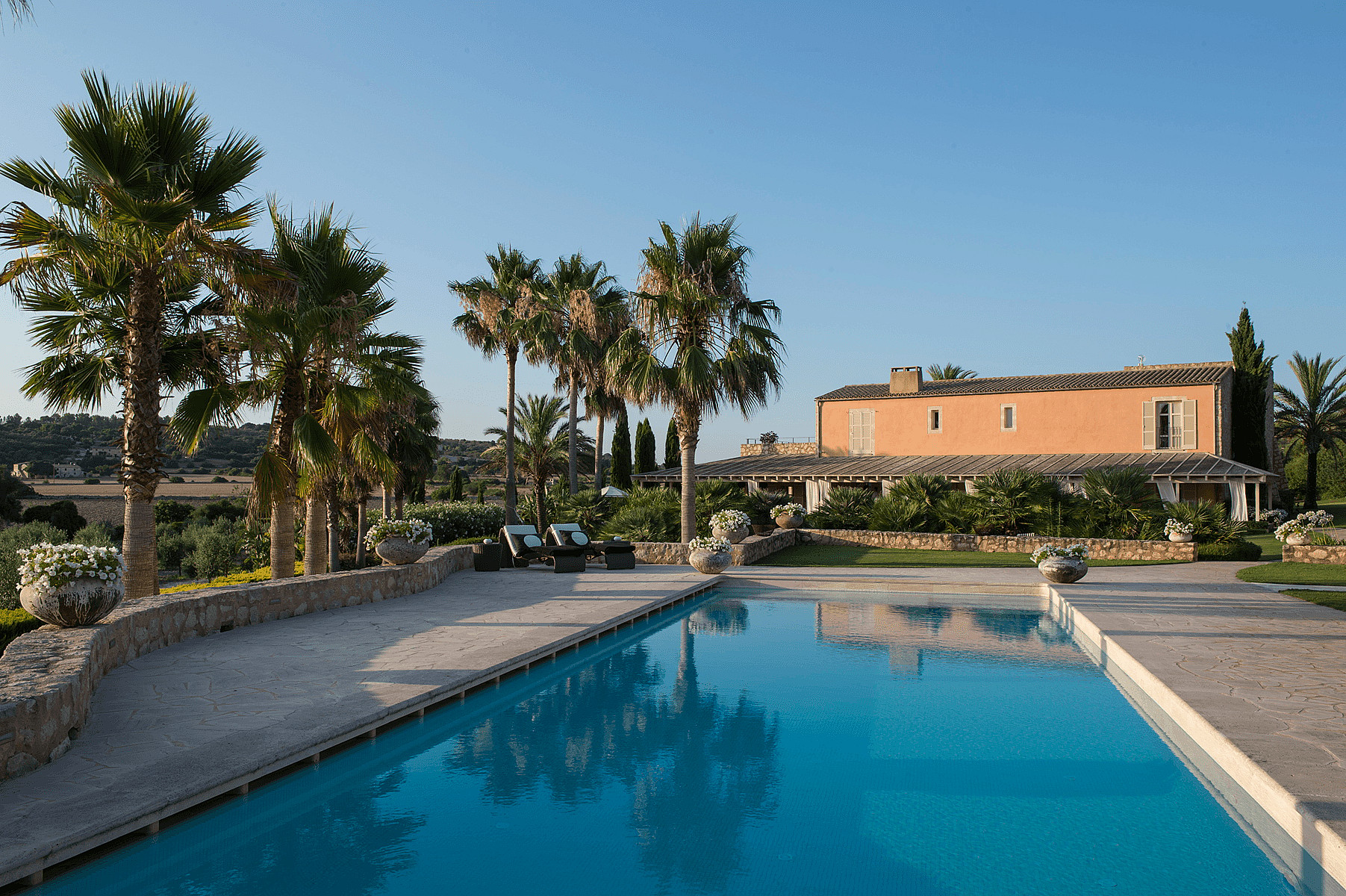 Rectangular swimming pool with blue water next to a stone-walled terrace, palm trees, and an orange farmhouse in Algaida.