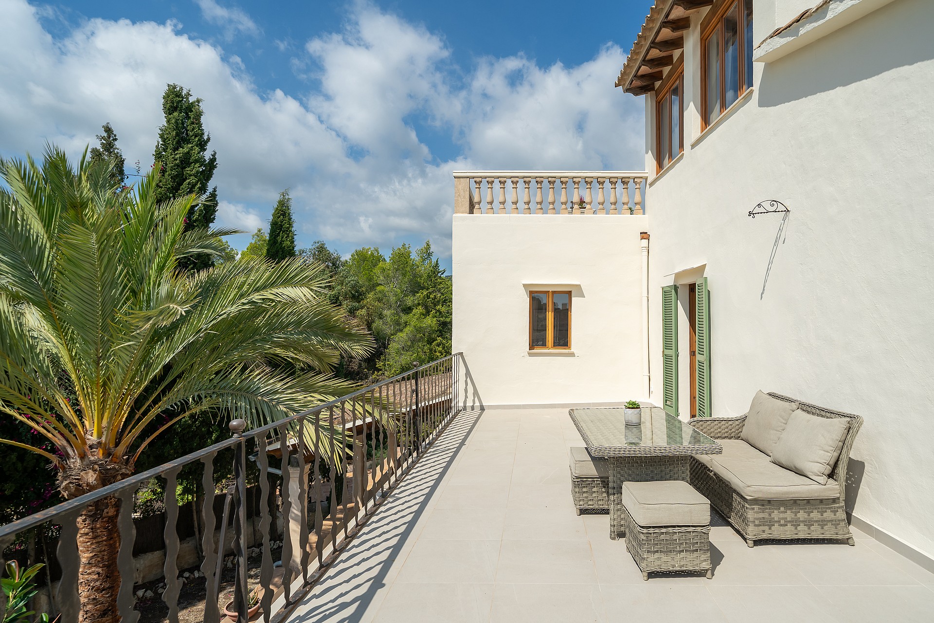 An elevated Mediterranean terrace featuring wicker furniture, wrought iron railings, and views of lush green trees.