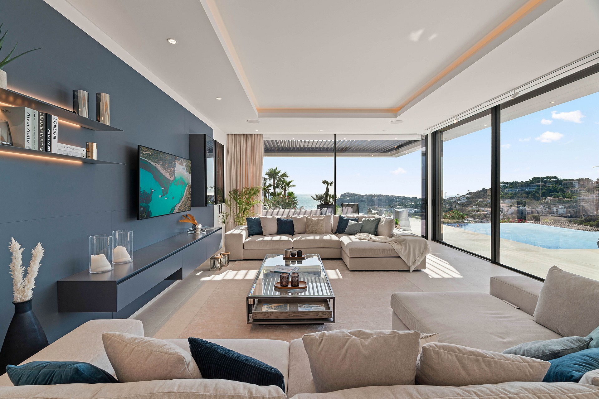 Spacious contemporary living room with a deep blue accent wall, neutral L-shaped sofas, and floor-to-ceiling glass sliding doors overlooking the sea.