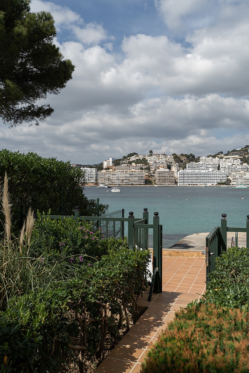 A view through a dark green garden gate over a terracotta terrace toward a calm turquoise bay and coastal buildings.