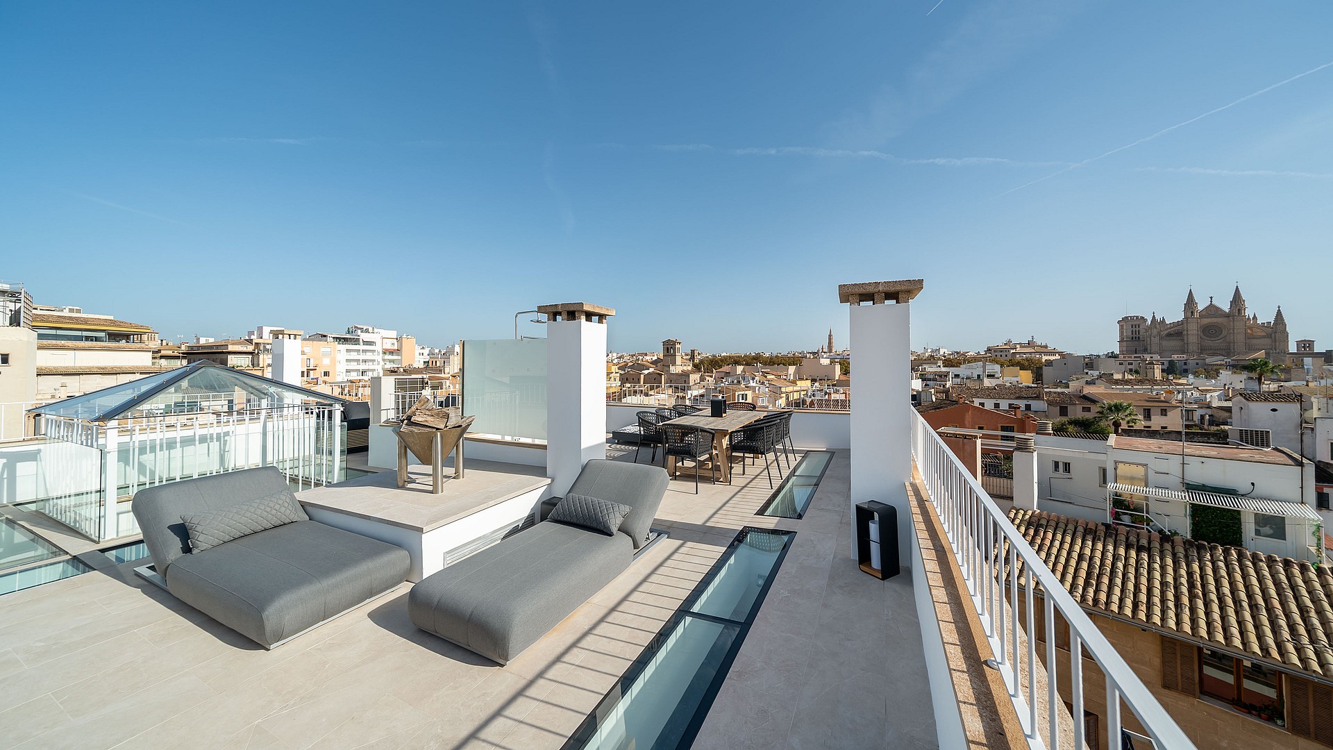Large rooftop terrace with grey loungers, glass skylights, and panoramic views of Palma Old Town and the Cathedral.
