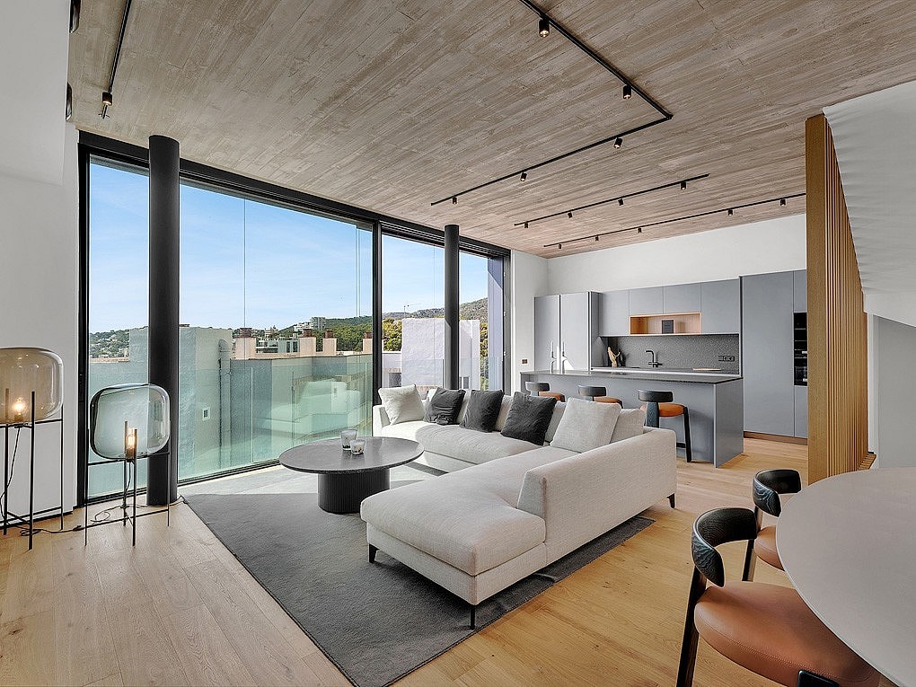 A modern open-plan living room with a grey sectional sofa, oak wood floors, and a textured concrete ceiling, leading to a glass-railed terrace with sea views.