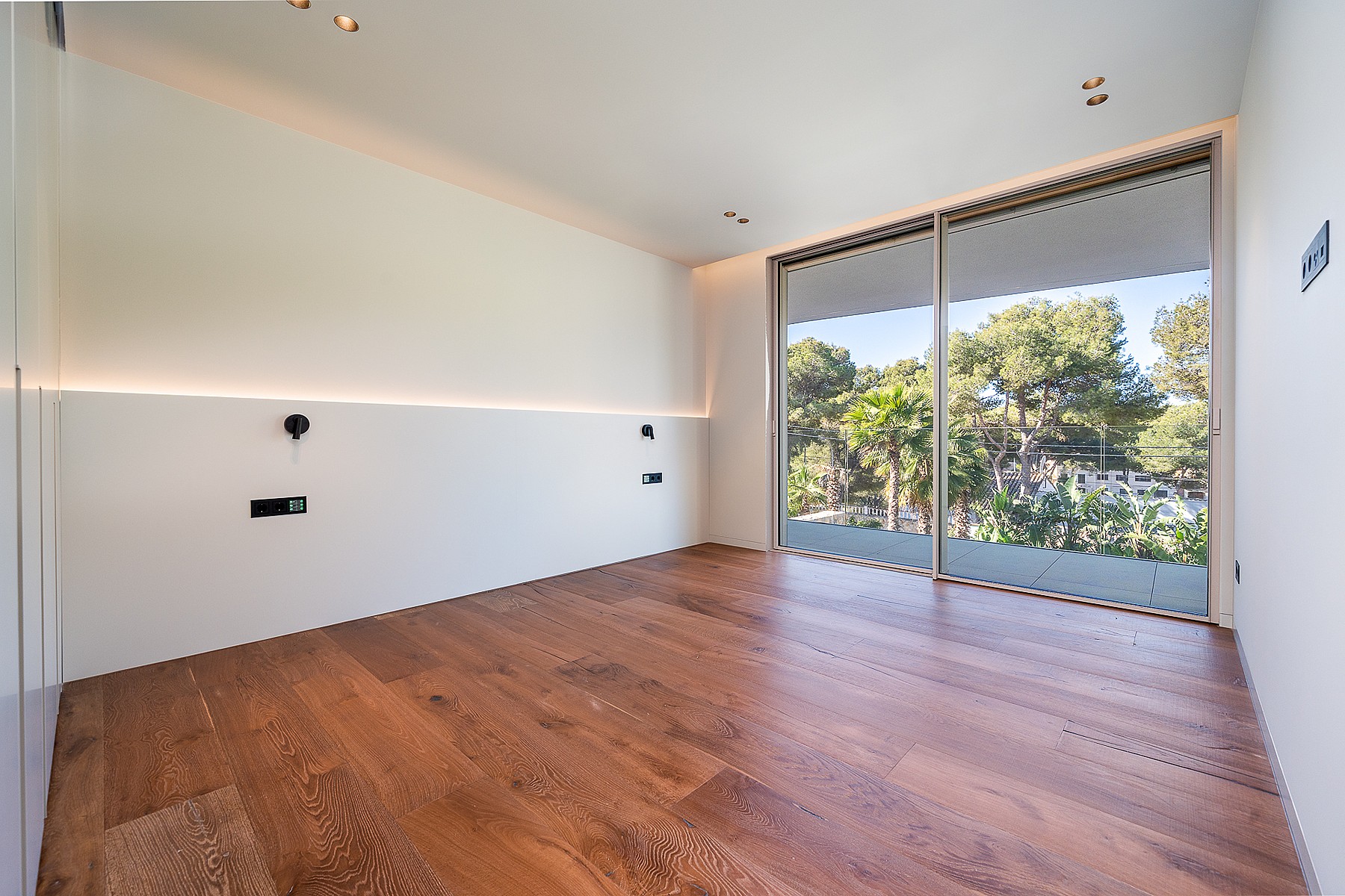 Empty bedroom with white walls, oak flooring, and sliding glass doors opening to a lush garden view.
