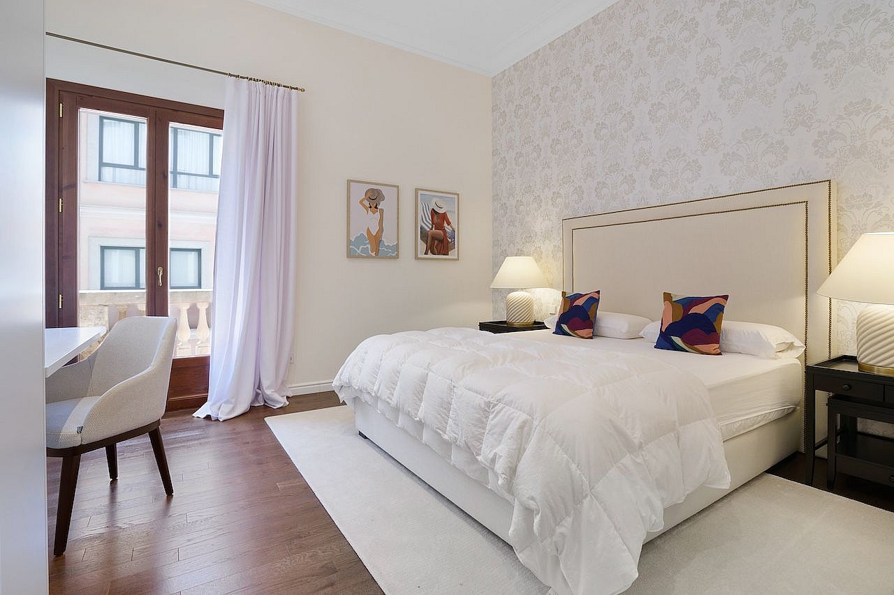 A bright bedroom with a king-sized bed, white linens, patterned wallpaper behind the headboard, and a balcony with white curtains.