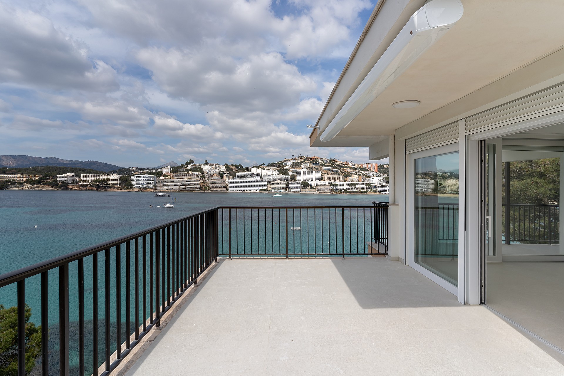 A spacious grey-tiled terrace with black metal railings faces a bay filled with boats under a cloudy sky.