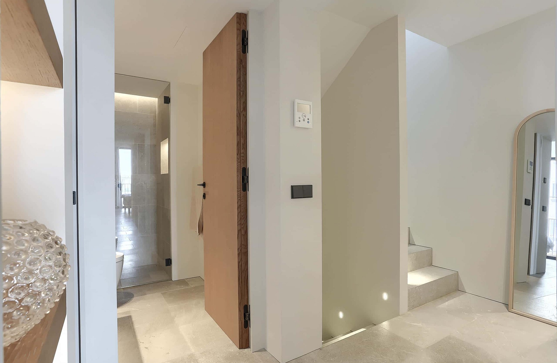 A bright, modern entrance hall featuring limestone flooring, a tall wooden door, white walls, and a recessed staircase.