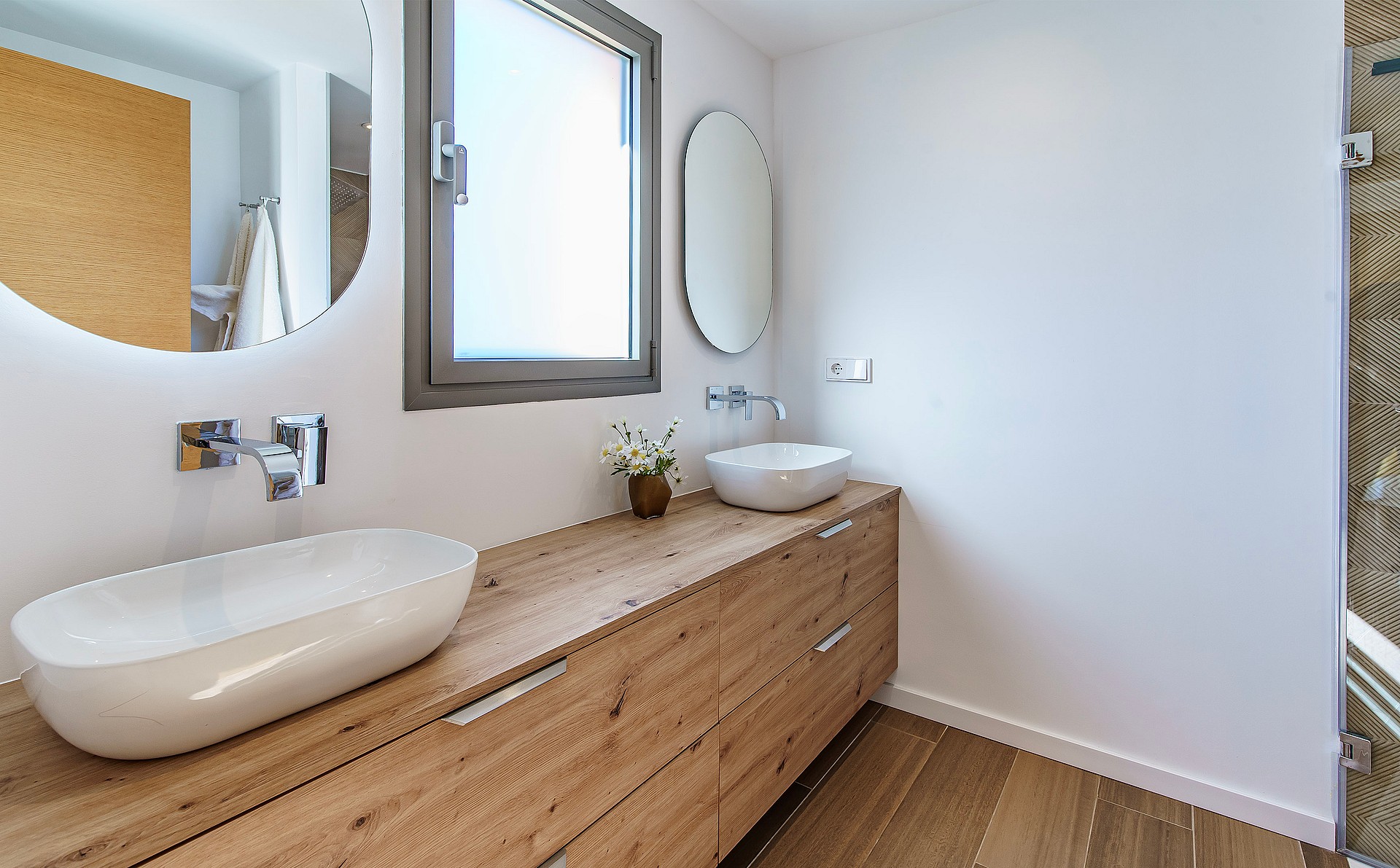Modern bathroom featuring a long wooden vanity with two white ceramic vessel sinks, circular mirrors, and wooden flooring.