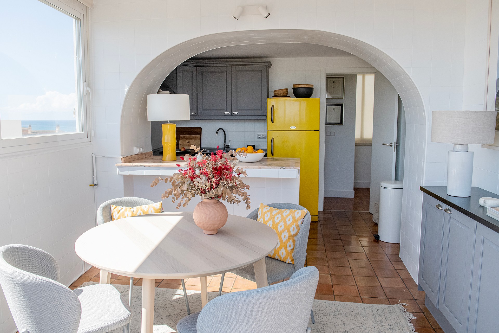 A small kitchen area with a yellow refrigerator, dark wooden cabinets, and a terracotta tiled floor seen through a wide arch.