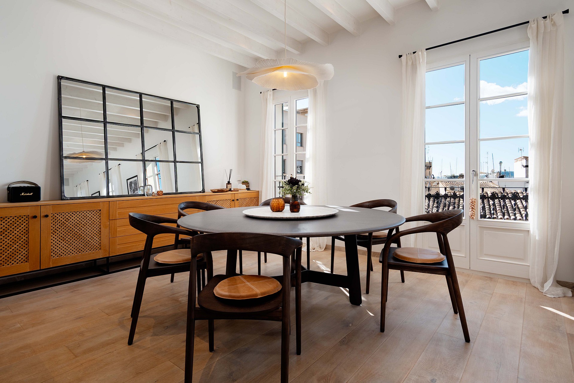 Round dark wood dining table with woven-seat chairs beneath a white beamed ceiling, French doors opening to rooftop views.