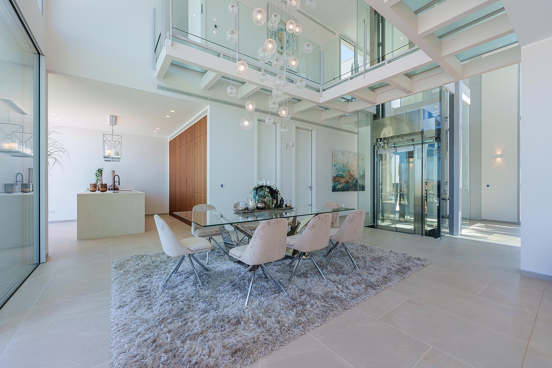 Dining room with a glass table, upholstered chairs on a textured rug, and a modern glass elevator in a double-height space.