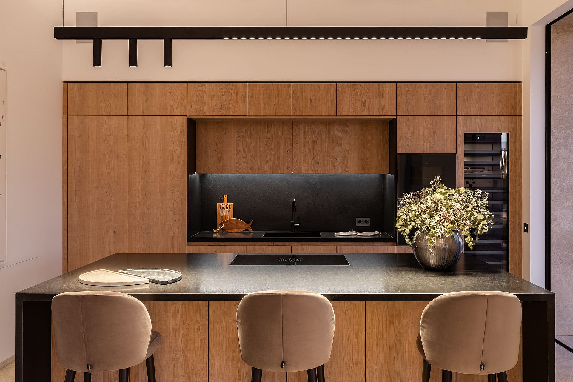 Modern kitchen featuring floor-to-ceiling oak cabinetry, a dark central island, and three beige upholstered stools.
