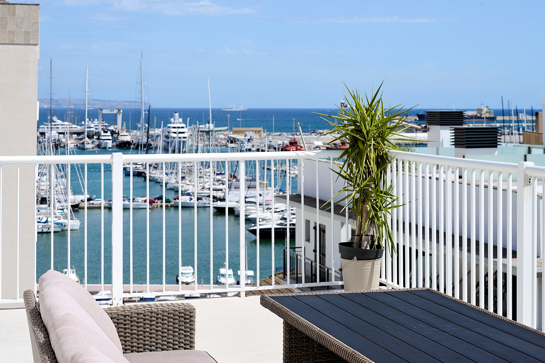 A private outdoor terrace with wicker seating and a potted plant, overlooking a marina filled with yachts.