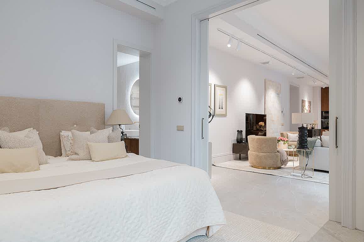 Interior view from a bedroom into an open-plan living room, featuring a beige headboard and neutral white decor.
