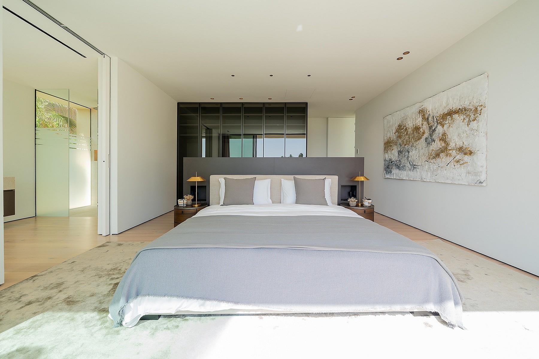 A minimalist master bedroom featuring a large bed, gray headboard, and abstract wall art, with an adjacent bathroom visible.