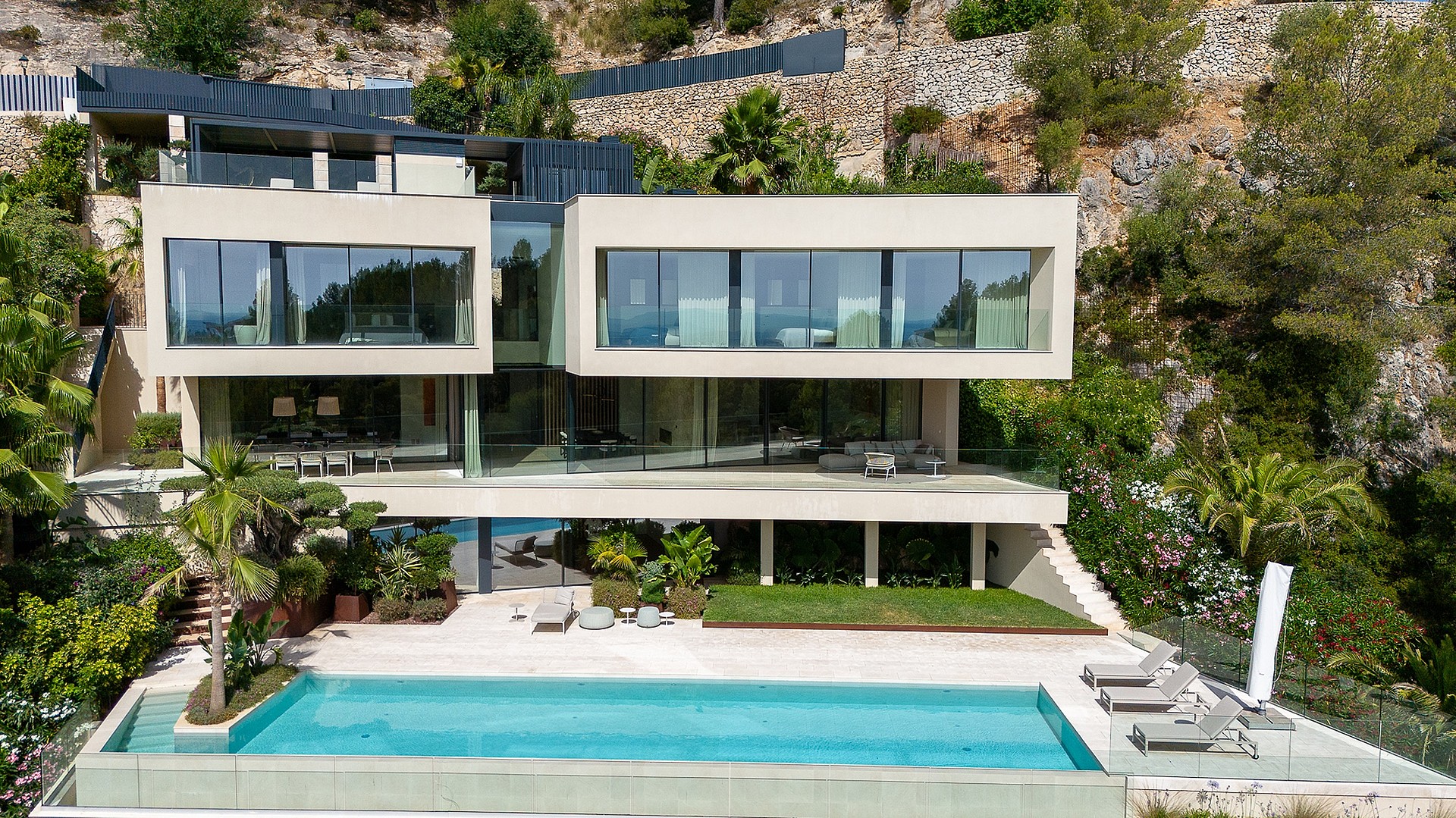 Exterior view of a multi-level contemporary villa built into a rocky hillside, featuring floor-to-ceiling glass walls, a rectangular swimming pool, and terraced gardens.