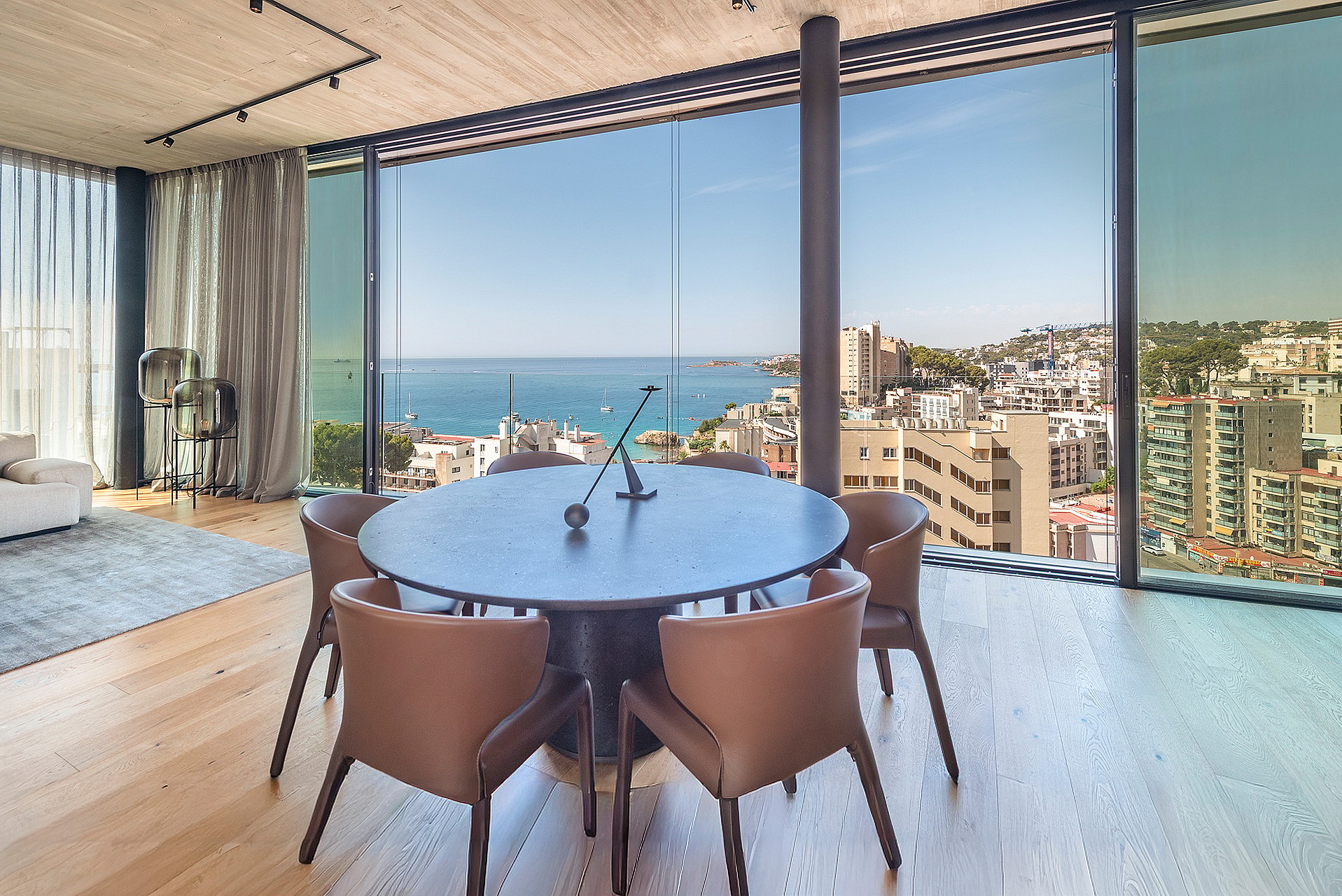 Round dark wood dining table surrounded by leather chairs next to floor-to-ceiling windows overlooking the sea and coastline.