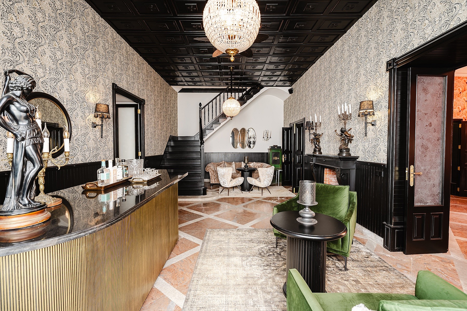 A spacious, eclectic interior featuring a large bar with metallic front, patterned wallpaper, black coffered ceiling, and a central staircase leading to the upper level.