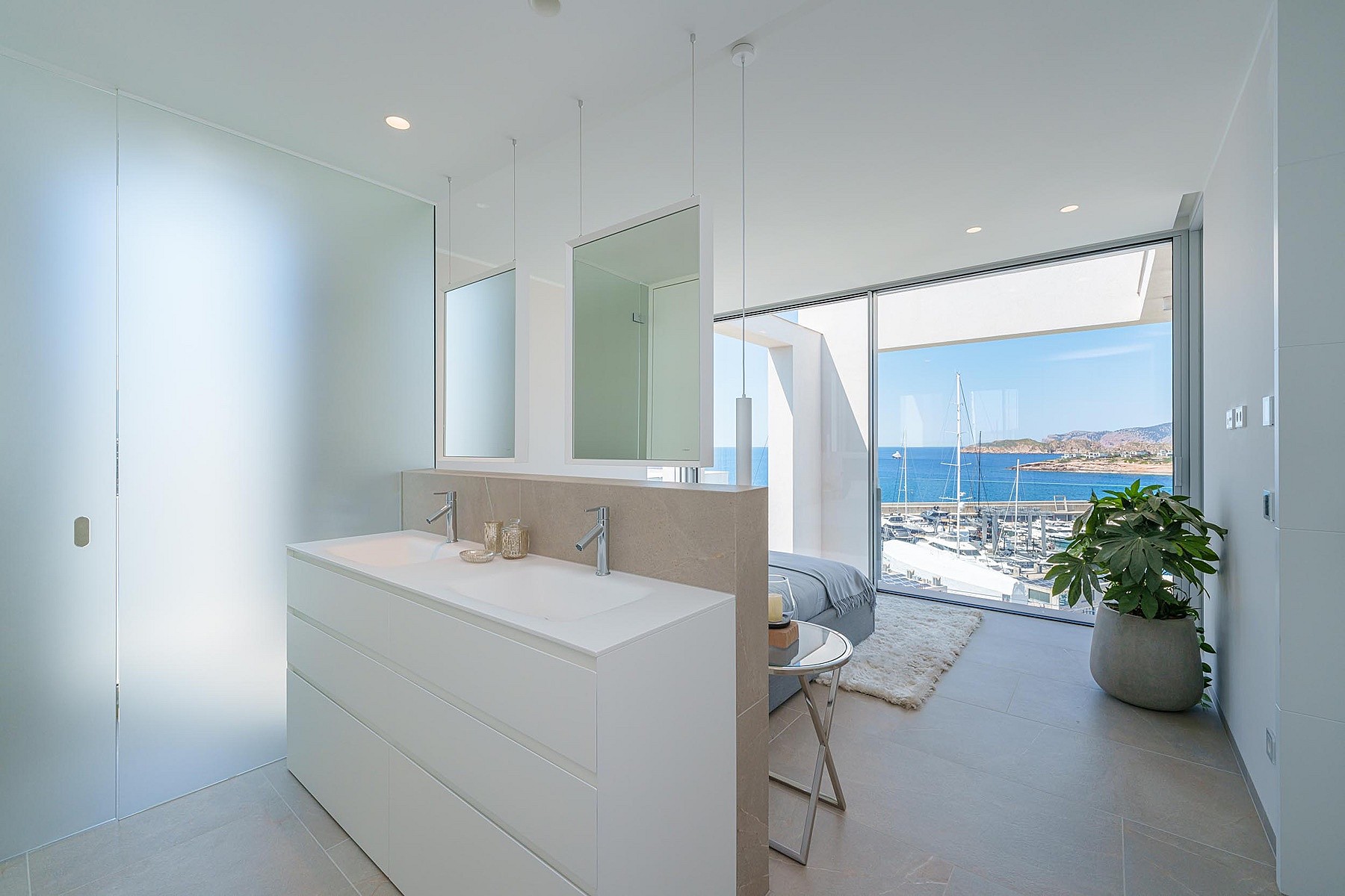A modern master bathroom with a double vanity island, floor-to-ceiling windows, and views over Port Adriano.