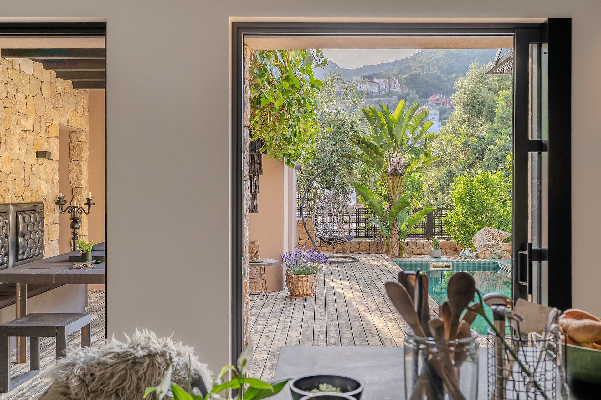Interior view from a kitchen counter looking out through an open glass door to a stone-paved terrace with lush greenery.