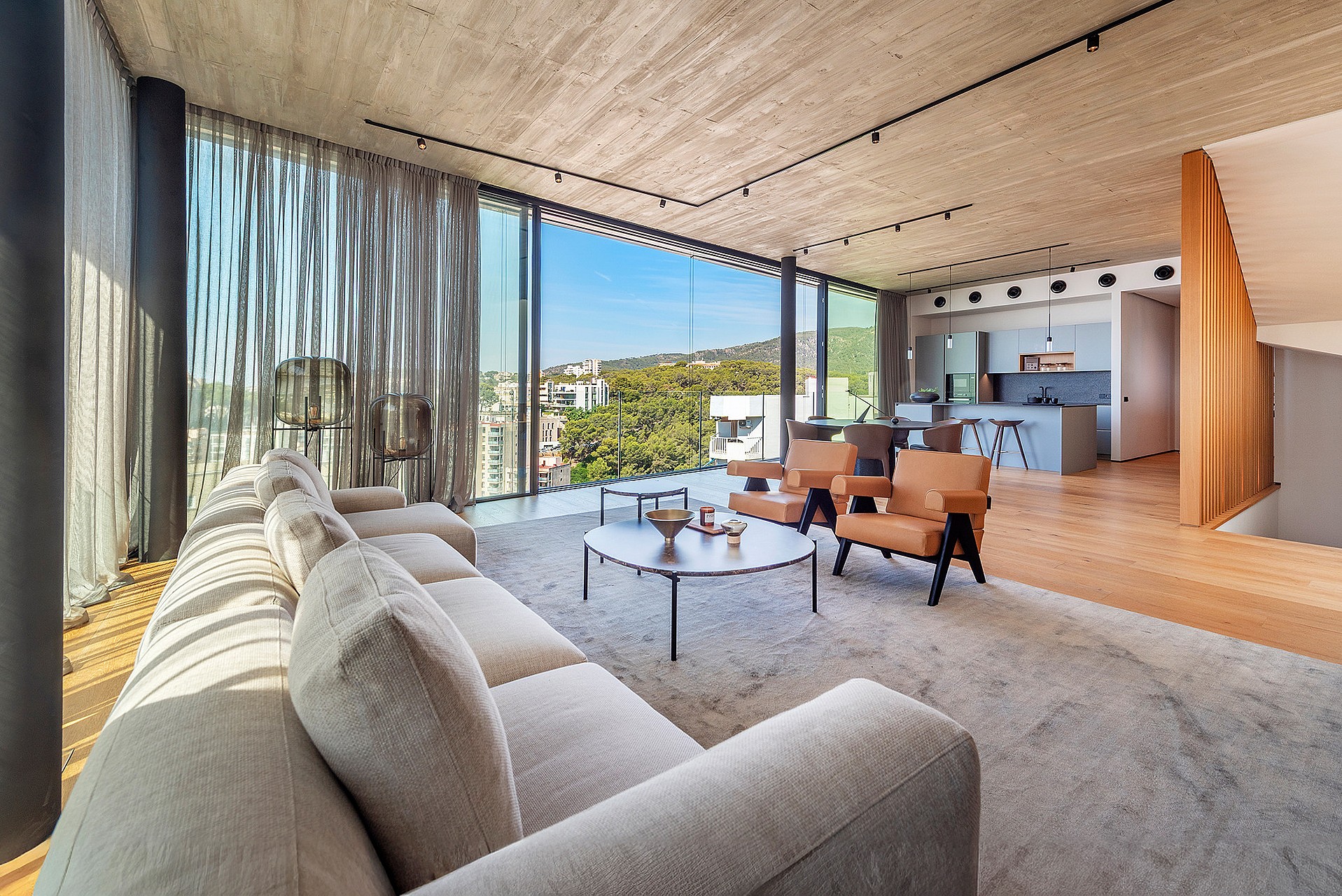 Wide-angle shot of a bright, open-plan living room with exposed concrete ceilings, light wood floors, and floor-to-ceiling glass walls looking out to coastal scenery.