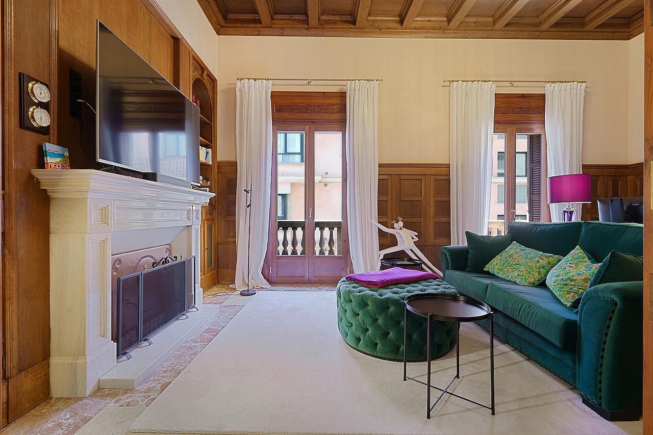 A formal living area featuring dark wood wall paneling, a stone fireplace, a green velvet sofa, and tall windows facing the city.