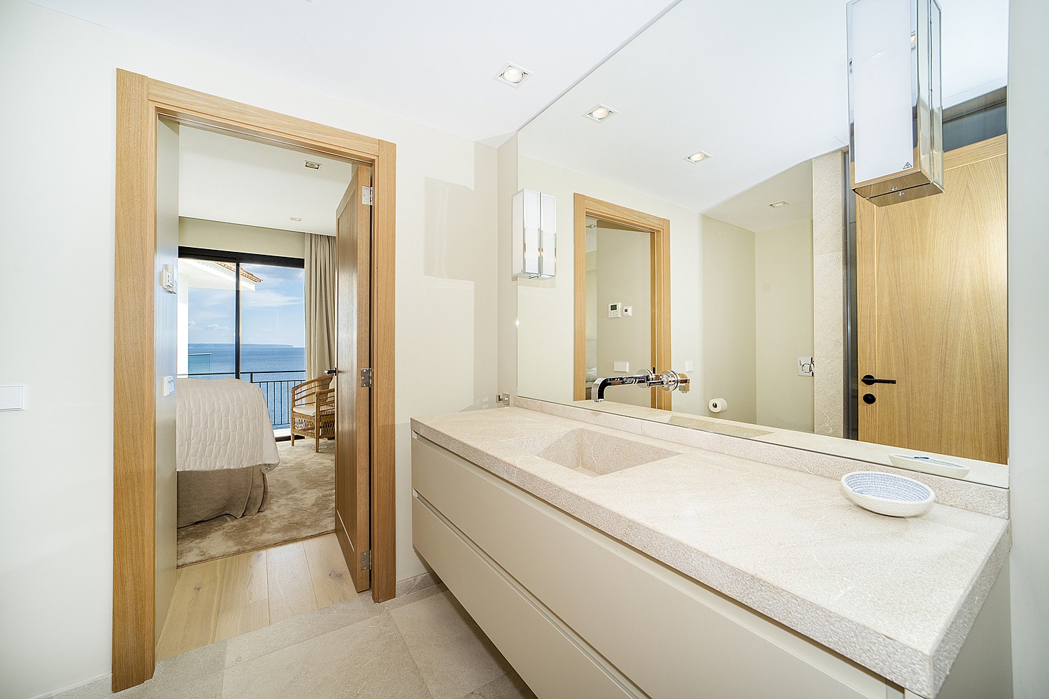 A modern bathroom vanity with a light stone countertop, a large frameless mirror, and a view through an open doorway into a bedroom overlooking the sea.