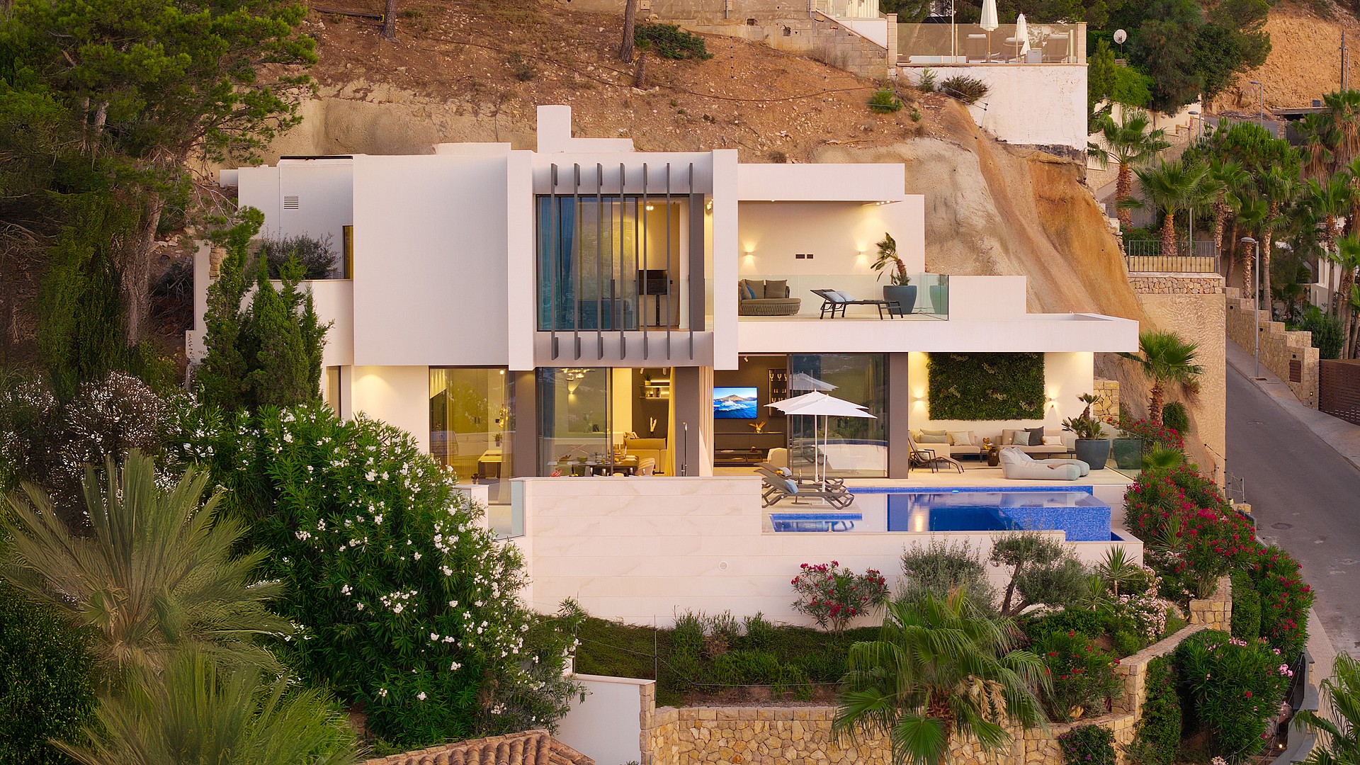 Exterior aerial view of a multi-level contemporary villa built into a rocky hillside with tiered terraces, pool, and lighting.