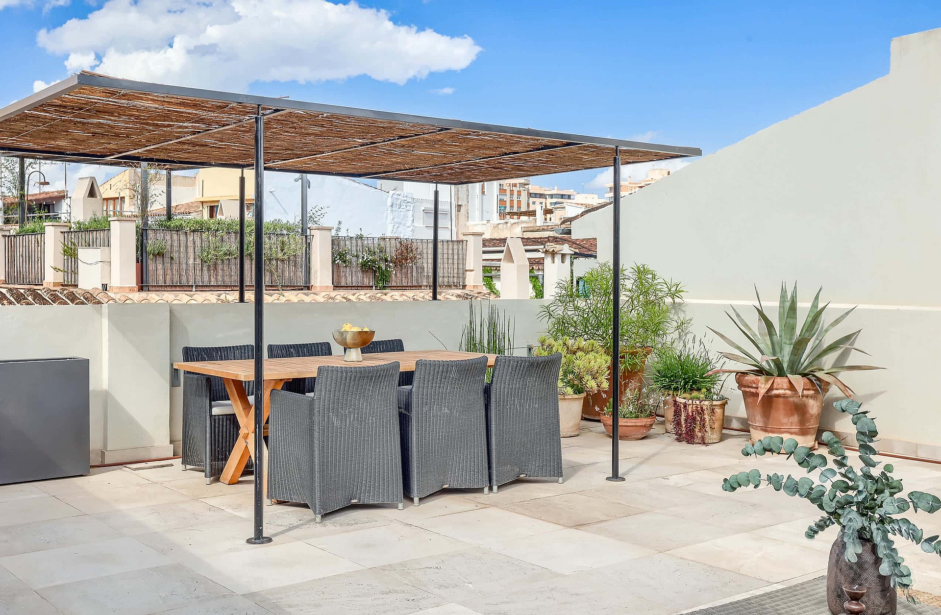 Rooftop terrace with a wooden dining table, woven chairs, and a metal pergola, surrounded by potted succulents and city views.