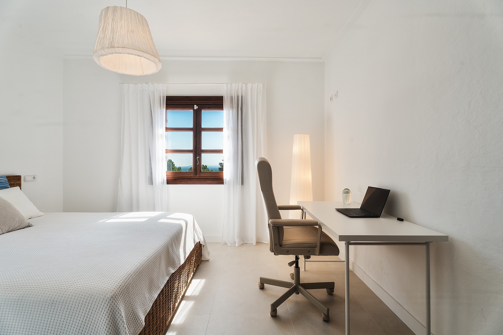 Dormitorio minimalista con cama, escritorio blanco con portátil y ventana hacia el paisaje montañoso.