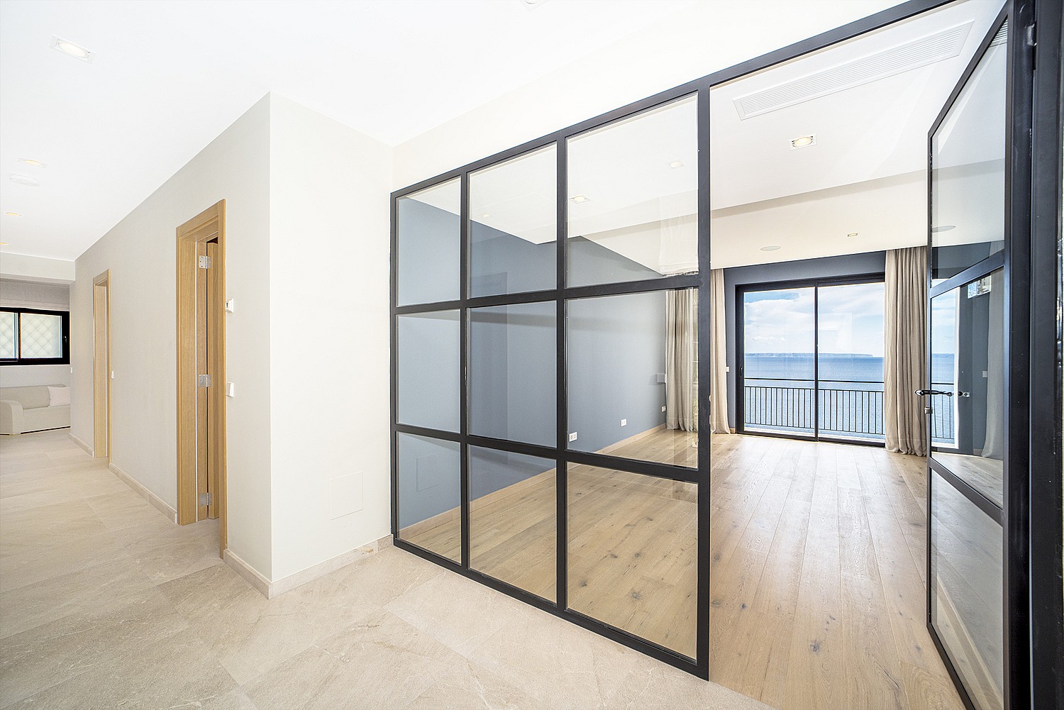 A walk-in wardrobe space featuring a black metal and glass partition, light oak wood flooring, and a view of the sea.