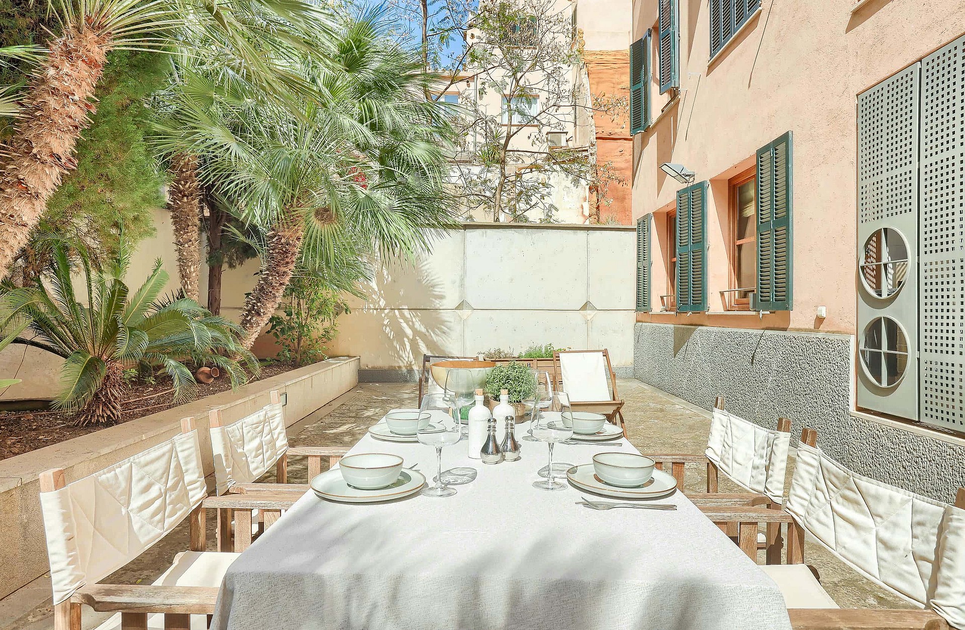 An outdoor dining table set with white linens beside a terracotta wall with green shutters, surrounded by palm trees.