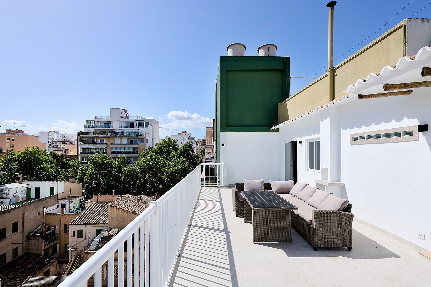 An elevated terrace with grey wicker seating and a dining table against a white exterior wall, overlooking the Santa Catalina neighborhood.