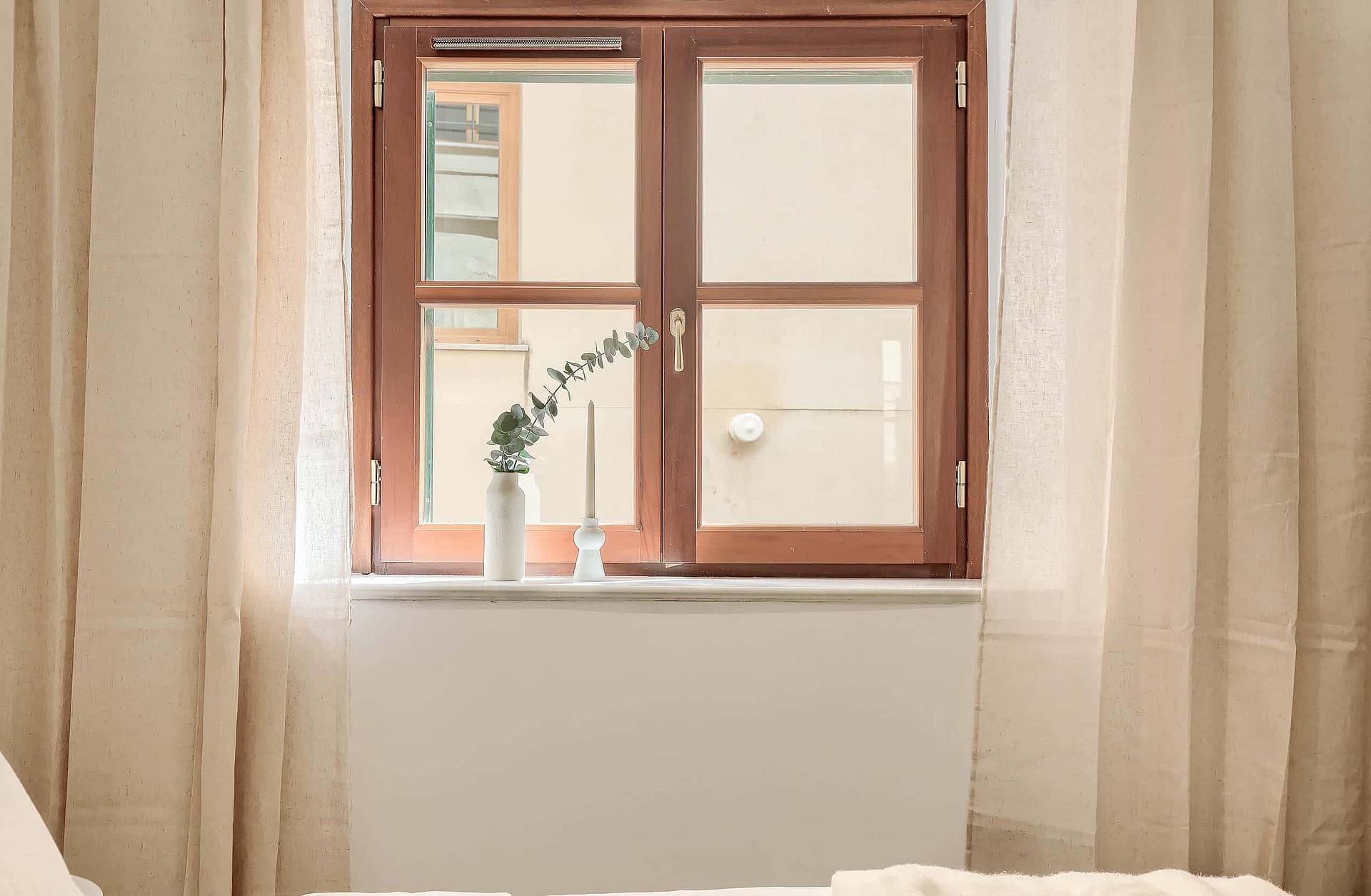 A close-up of a wooden window frame looking out onto a residential building facade, framed by floor-length linen curtains.