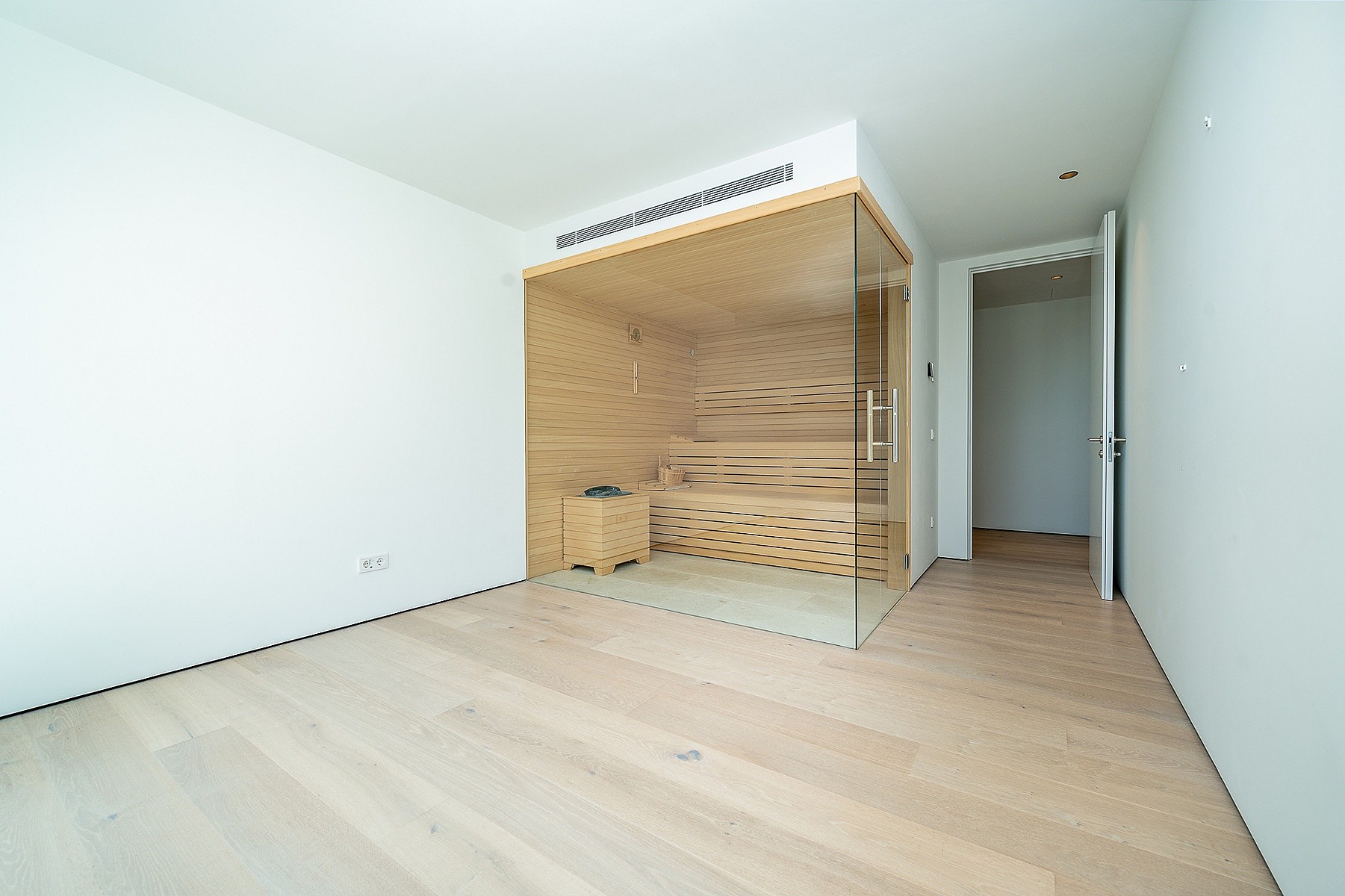 Modern home sauna with light wood paneling and glass doors inside a minimalist white room with light oak flooring.
