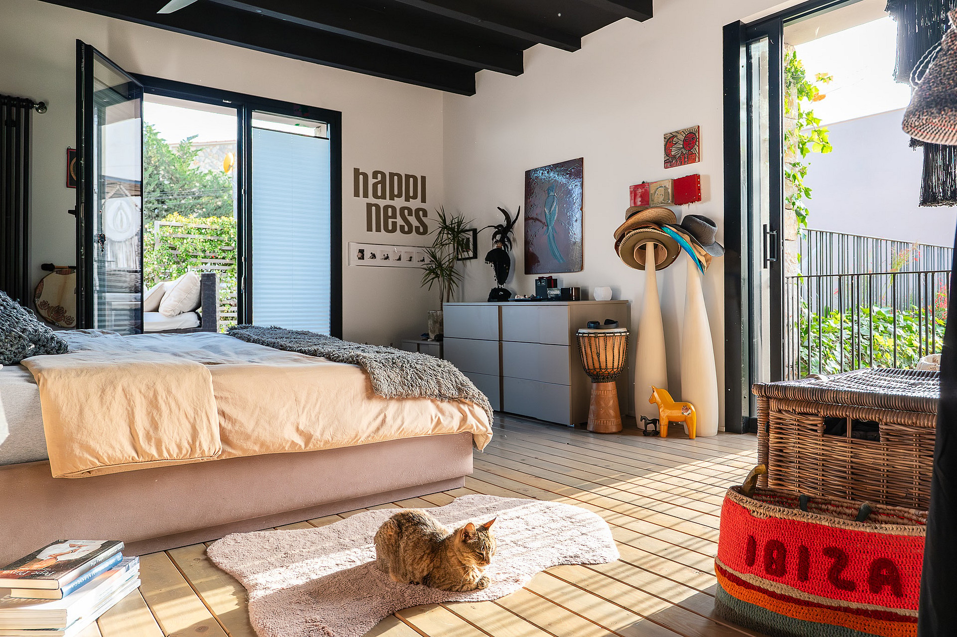 Interior of a bedroom featuring a platform bed, wooden floorboards, and glass doors opening to a garden, with eclectic decor.