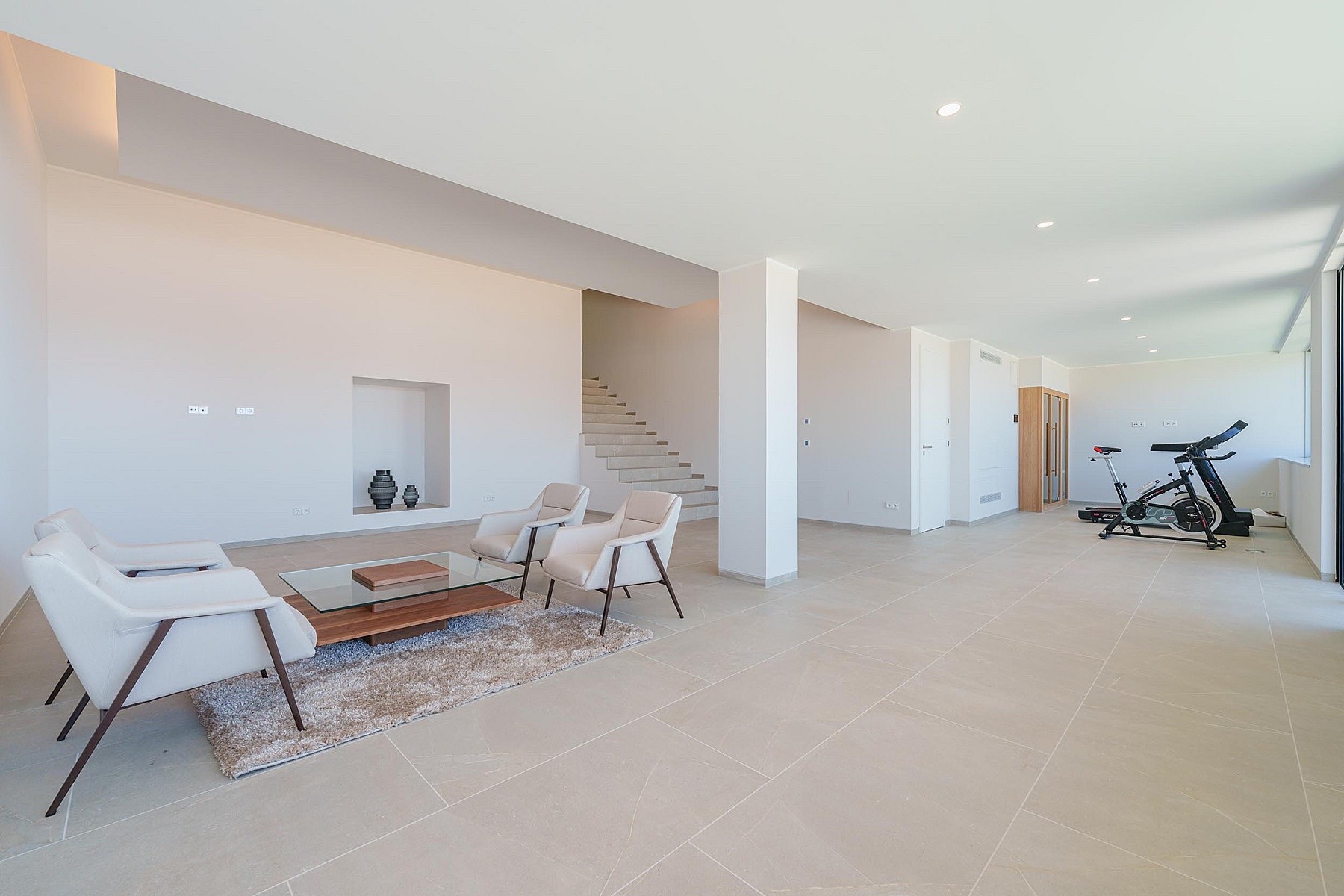 Spacious open-concept room with white walls, beige stone floors, a small lounge area, and exercise equipment in the background.