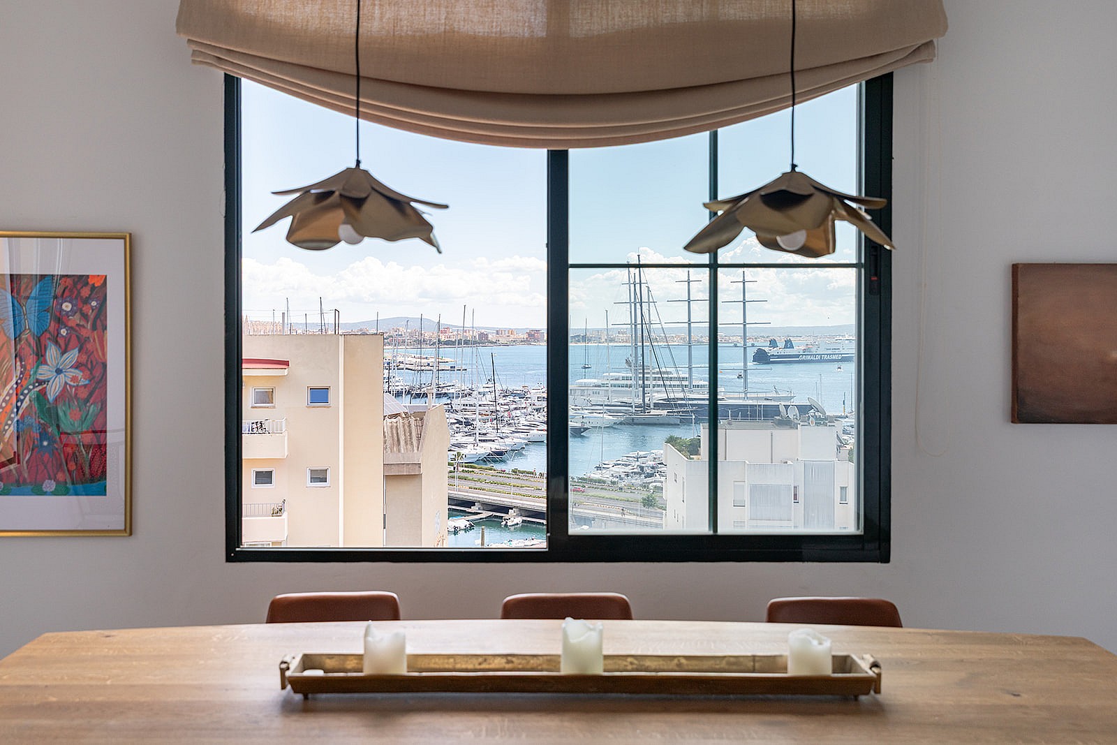 A dining table centered in front of a window looking over a harbor with boats, featuring pendant lights and a wooden tray.