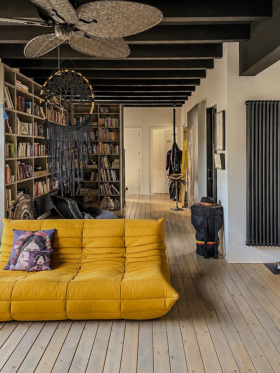 A living room interior featuring a bright yellow tufted sofa on pale wooden floorboards, dark exposed ceiling beams, and a floor-to-ceiling bookshelf.