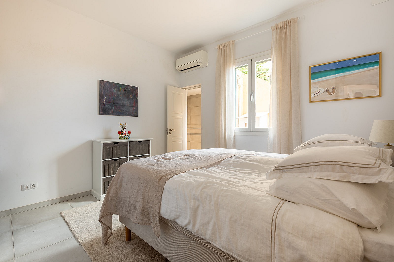 A bright bedroom with a white bed, beige throw, textured rug, and white walls. A dresser and wall art are visible.