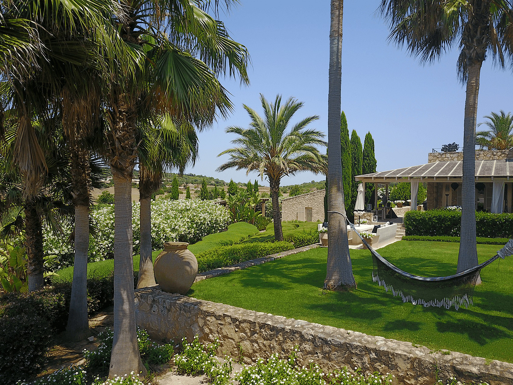 A wide view of a private garden featuring a manicured lawn, several tall palm trees, a hammock suspended between trees, and a low stone wall in the foreground.