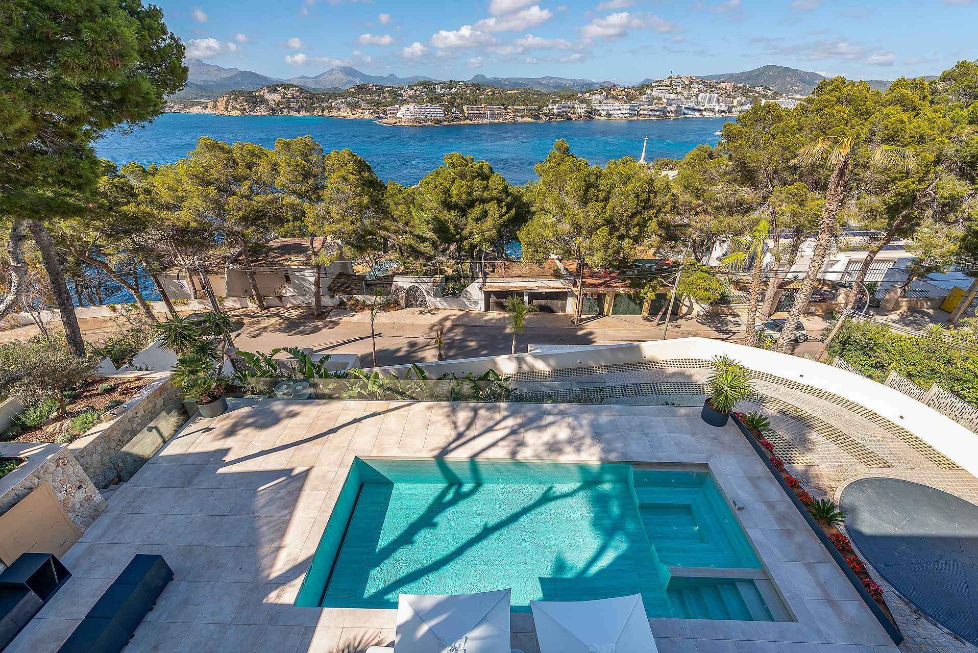 High-angle view of a rectangular swimming pool with turquoise water, set in a stone-paved terrace overlooking a bay.