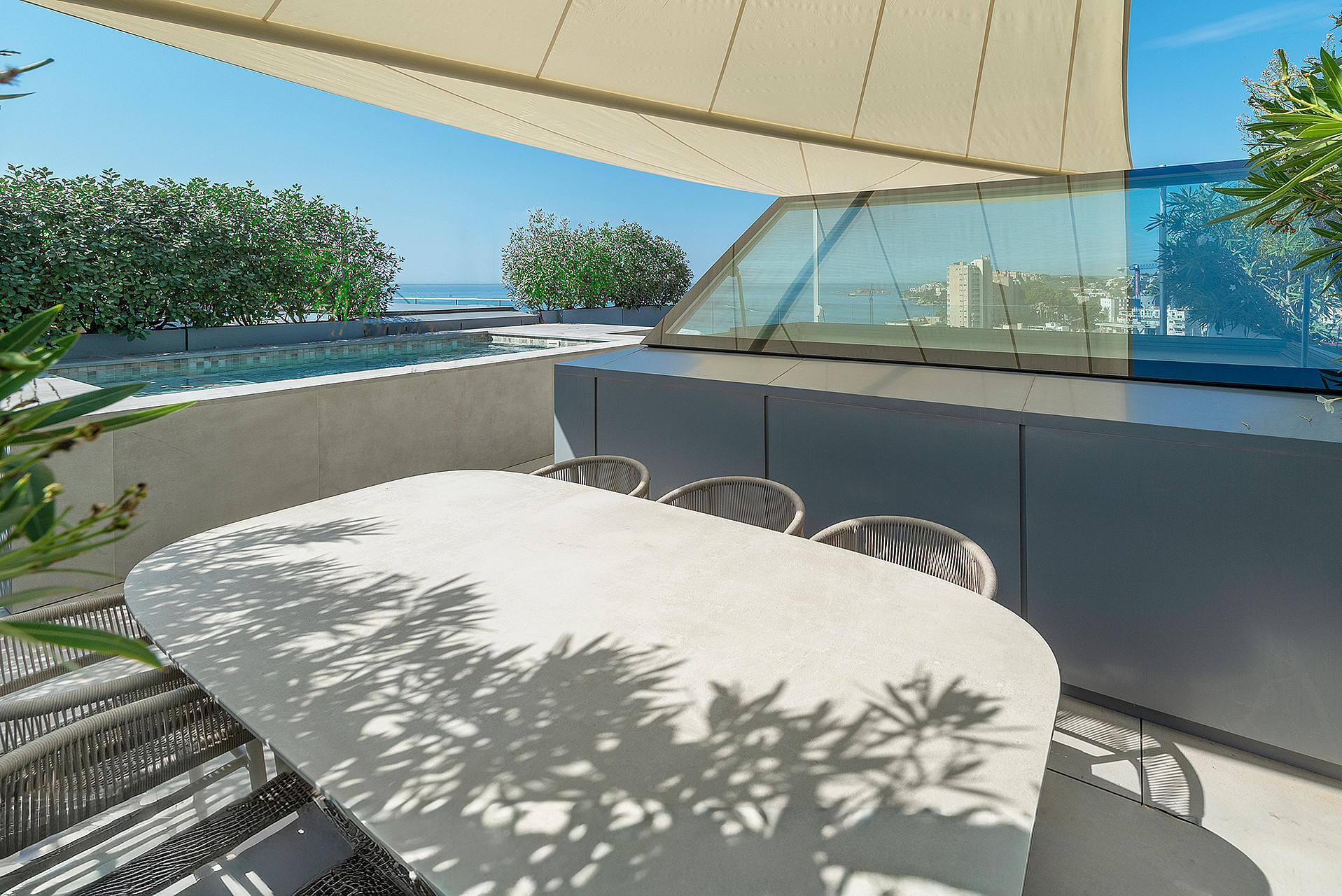 An outdoor dining table with woven chairs on a sunny rooftop terrace, featuring a glass partition and sea views.