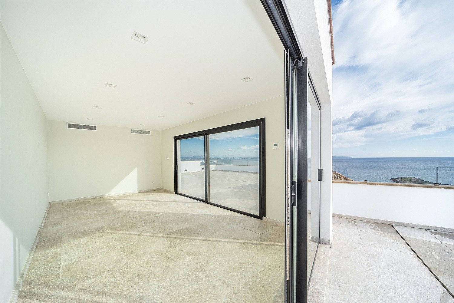 An empty modern room with light stone flooring and large sliding glass doors opening onto an outdoor sea-view terrace.