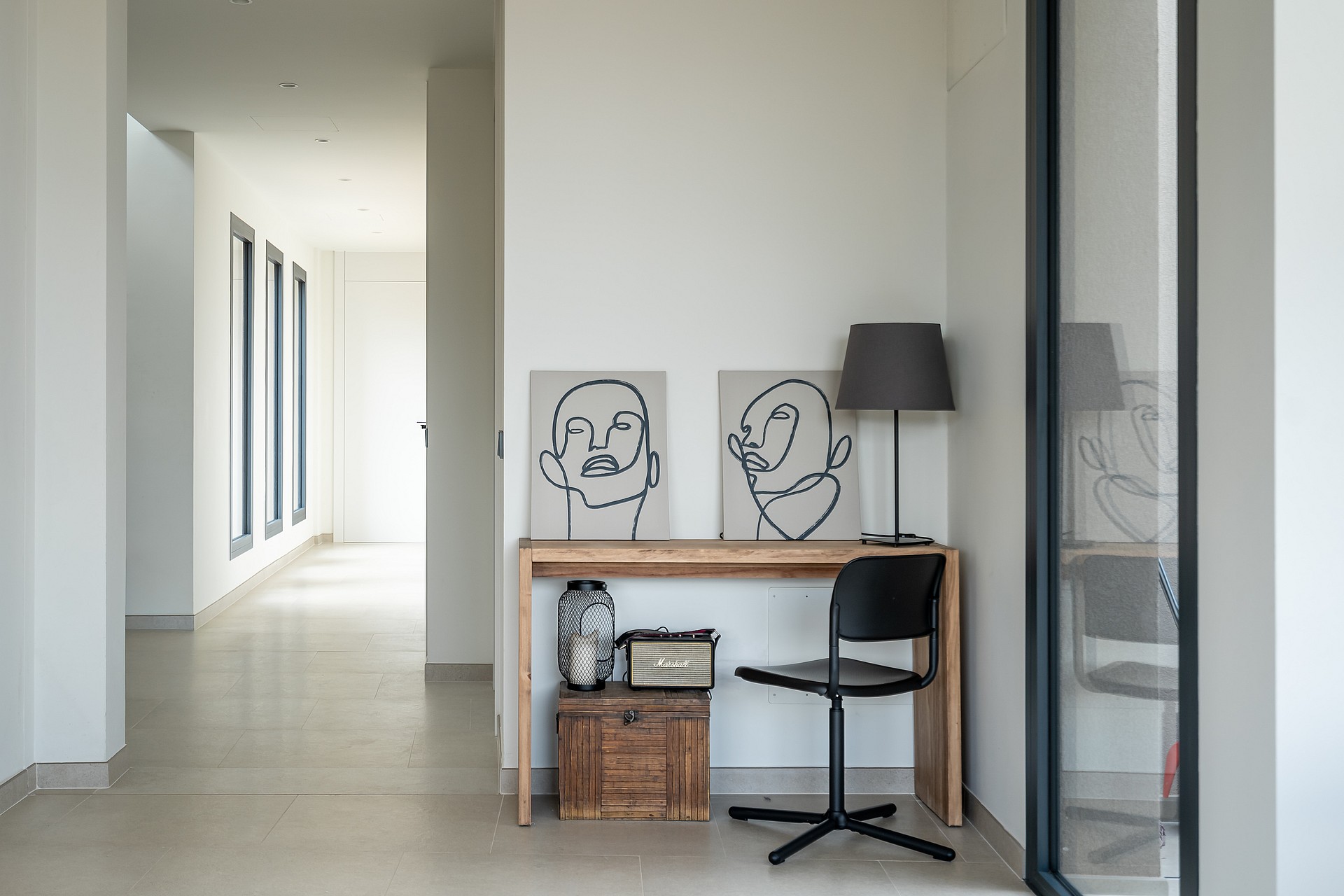 A minimalist hallway with a wooden desk and a black chair, featuring abstract line art prints against a white wall.