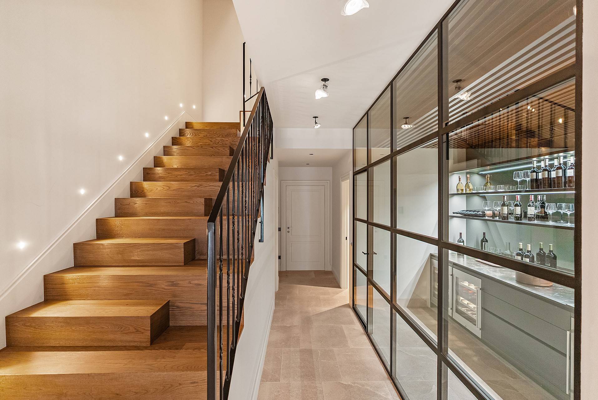 Contemporary hallway featuring a black steel-framed glass wine cellar next to a wooden staircase with iron railings.