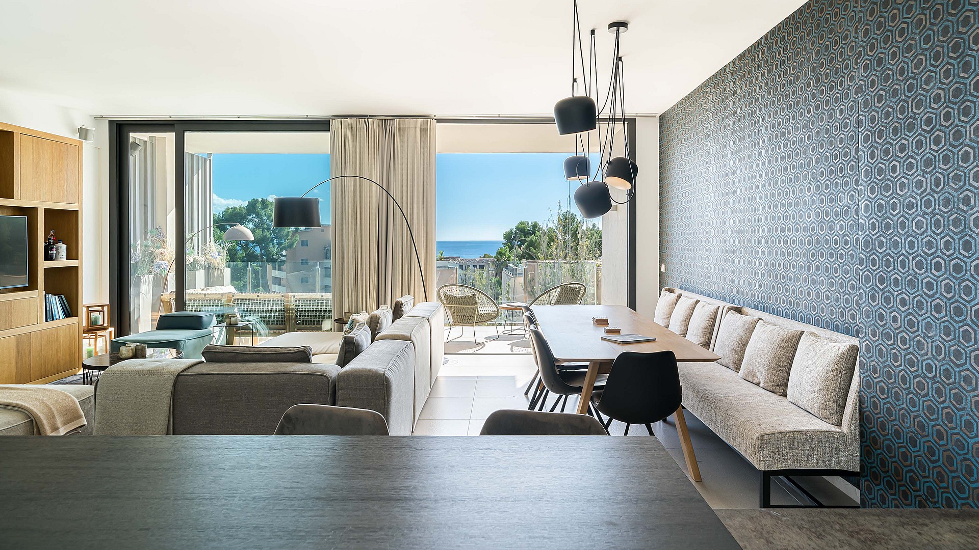 Open-plan living and dining room with a long beige bench seat, dark pendant lights, and floor-to-ceiling balcony views.