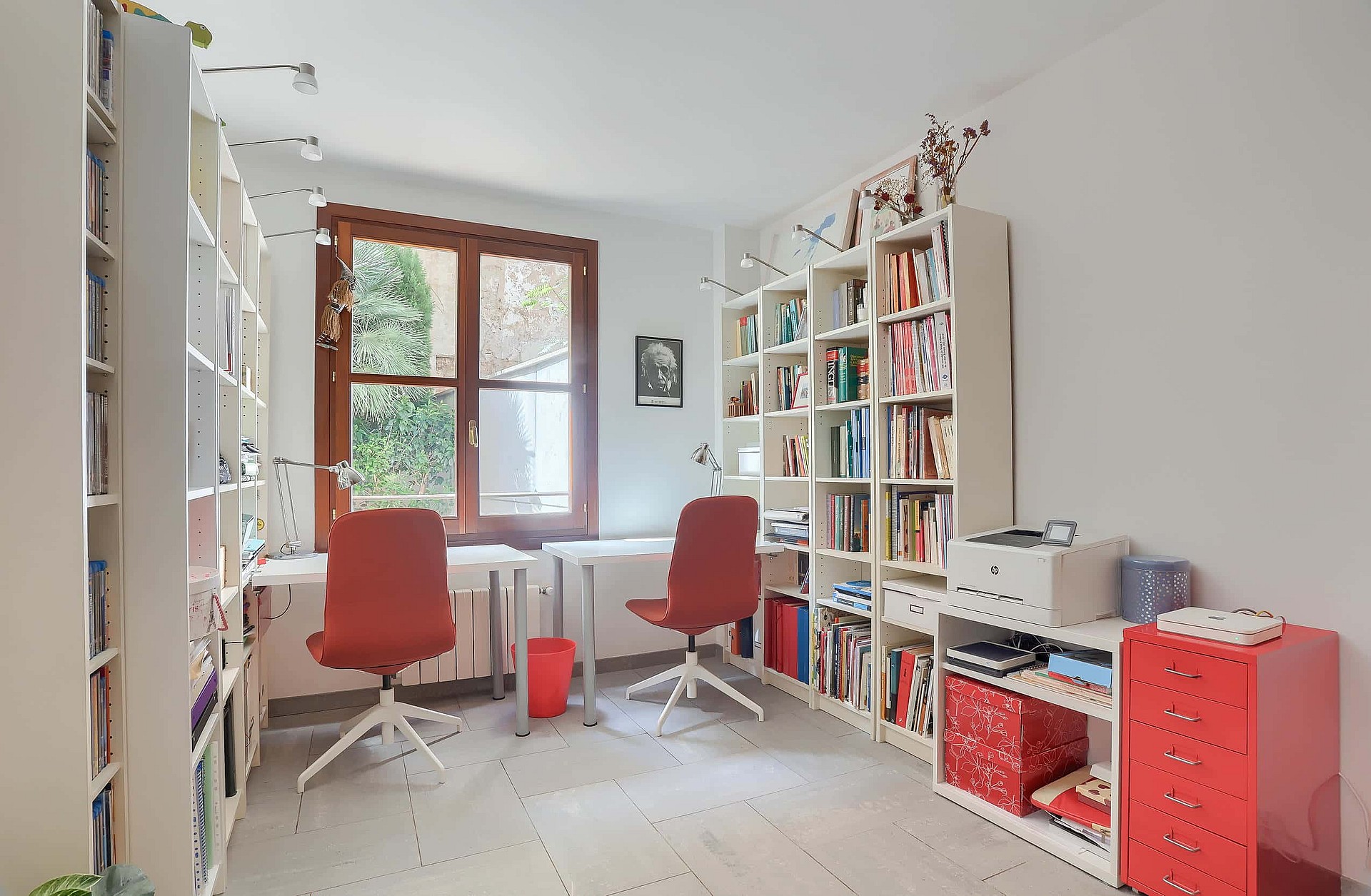 A home office featuring two white desks, red rolling chairs, and floor-to-ceiling white shelving units under a wooden window.