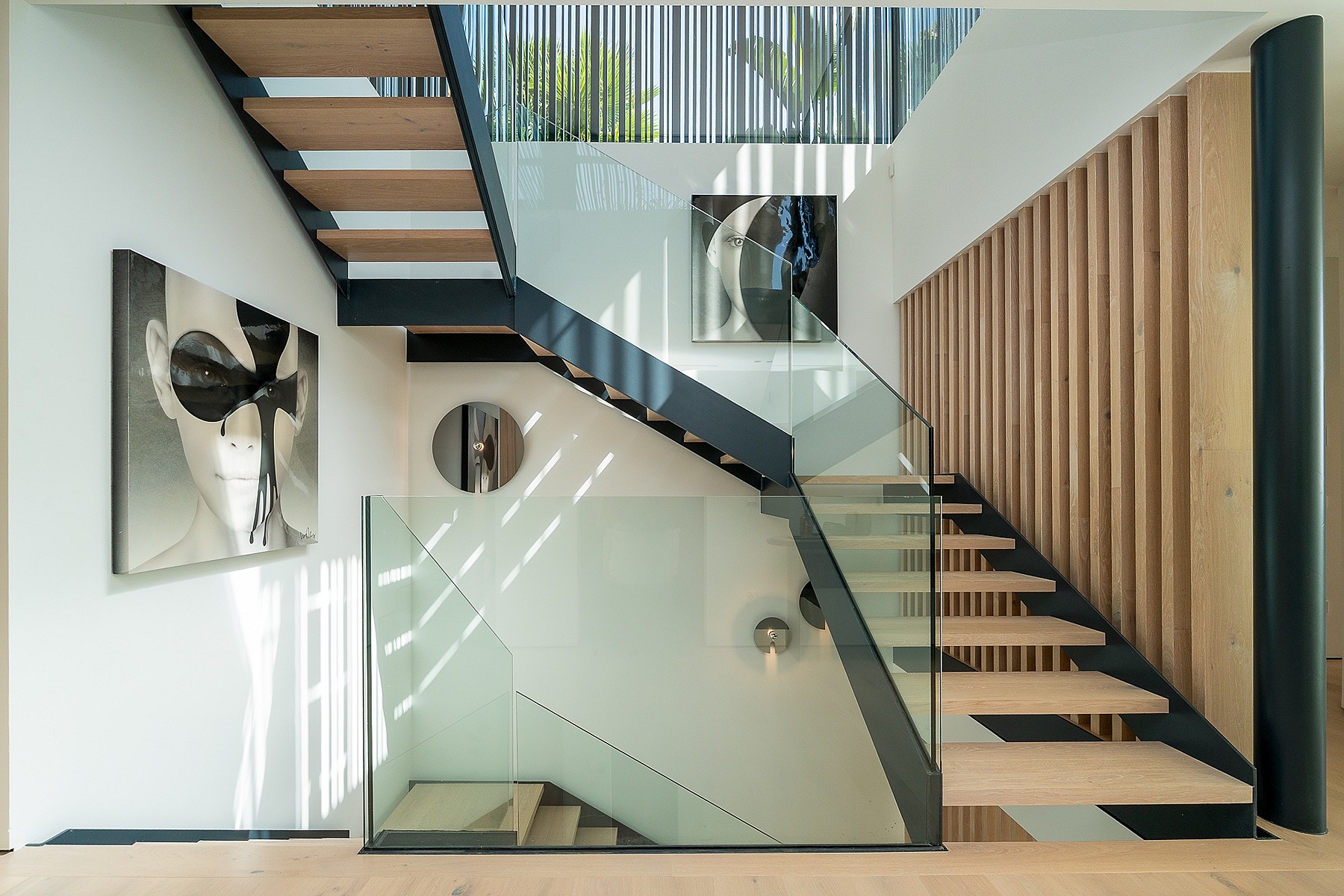 Modern open-tread staircase made of light wood and black metal, featuring frameless glass railings and vertical wooden slats.